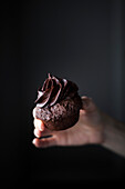 a hand holding a chocolate cupcake with chocolate frosting.