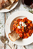 A bowl of sausage goulash topped with soured cream and served with crusty bread and red wine
