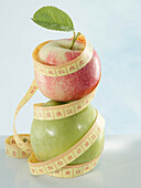 A red apple with a leaf on a green apple wrapped with a tape measure