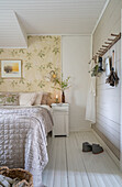 Doppelbett mit silberfarbener Tagesdecke und Tapete mit Blütenmotiv im Schlafzimmer
