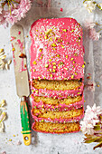 A part-sliced loaf cake with pink icing topped with coconut and pistachio nuts