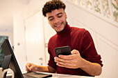 Young man with smart phone working from home on laptop