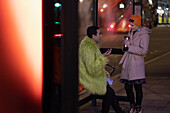 Couple with coffee talking at bus stop in city at night