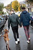 Gay male couple holding hands walking dog on wet street