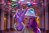 Woman in fedora gesturing peace sign under neon