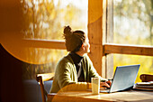 Woman working on laptop looking out window in sunny cafe