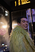Happy eccentric young man in feather coat at city bus stop