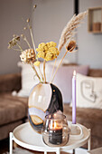 Glasvase mit Trockenblumen, Kerze und Windlicht auf Beistelltisch