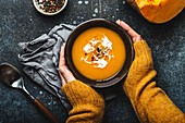 Female hands in yellow knitted sweater holding a bowl with pumpkin cream soup