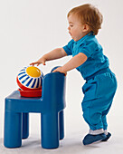 Toddler standing leaning over chairback to reach a ball