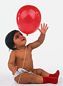 Baby wearing red socks and nappy, reaching for a red balloon