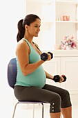 Smiling pregnant woman sitting on chair lifting weights