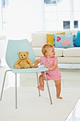 Baby girl standing using a chair with teddy bear on the seat