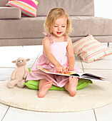 Girl sitting reading a book, pointing at the page with toy lamb beside her