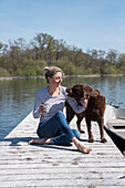 Blonde Frau sitzt mit Hund auf Holzsteg