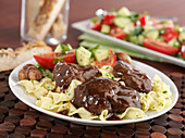 Braised beef short ribs with braising liquid on a bed of pasta with cucumber and tomato salad