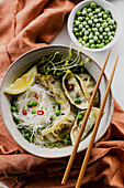 Gyoza, dumpling soup with thin rice noodles, broth, peas, chilli, sprouts and a slice of lemon, orange linen and chopsticks