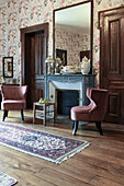 Upholstered chairs in front of fireplace with mirror above in room with floral wallpaper