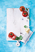White wooden board, tomatoes, basil, and a cheese grater on blue background
