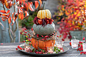 Pumpkin tower as table decoration: Pumpkins 'Sunny Hokkaido', 'Hungarian Blue' and 'Sweet Dumpling' with wreaths of rose hips, leaves and lanterns placed on top of each other on a bowl with lanterns, snowberries, rose hips and ornamental apples.