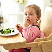 Baby girl sitting in high chair eating broccoli