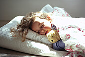 Young girl lying tucked up in bed asleep with teddy bear