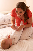 Mother putting babygro on baby who is lying on bed