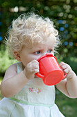 Baby girl in a garden drinking from red plastic beaker