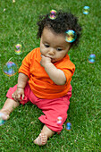 Baby girl sitting on lawn in garden surrounded by bubbles