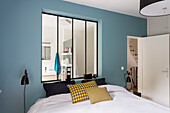 Doppelbett vor Innenfenster mit Blick ins Bad im Schlafzimmer mit blauer Wand