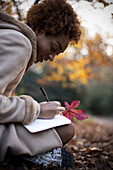 Young woman with autumn leaf writing in journal in park