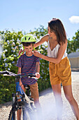 Mother helping son with bike helmet in sunny driveway