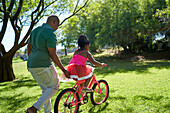 Father pushing daughter on bicycle in sunny summer park