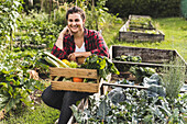 Lächelnde junge Frau sitzt mit Gemüse in Kiste auf Hochbeet im Gemeinschaftsgarten