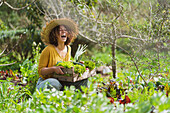 Lachende Frau, die eine Kiste mit grünem Gemüse hält, während sie im Garten hockt