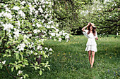 Junge Frau mit weißem Kleid und Schlapphut, die barfuß im Garten mit blühenden Apfelbäumen läuft