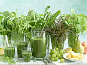 Green smoothies made of different kinds of leaf mustard, in front of grapes, pineapple, pear, apple