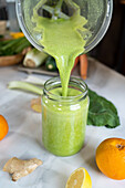Pouring tasty smoothie from blender bowl into jar at table with citrus fruits at home