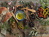 Still life with green and black olives on wooden spoons, dried tomatoes, pickled chilies, different herbs and bread