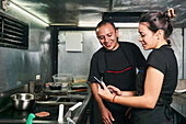 Content female worker showing mobile phone to smiling guy while standing in food truck with frying burger patty on stove