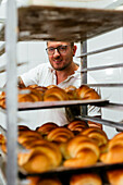 Male baker with tattoos on arm baking croissants in large metal oven during work in bakery