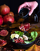 Adding olive oil in bowl with appetizing vegetarian pomegranate salad served on table with autumn leaves and cones
