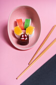 Colorful sweet sushi served on ceramic plate with wooden chopsticks placed on pink and black background
