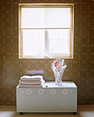 Modern bathroom details of a pile of towels and a glass vase full of shells on top of a decorated bathroom chest square window retro pattern tiles