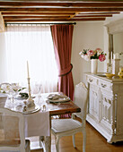 A traditional country dining room with exposed wooden beams on the ceiling above a vintage buffet next to a wooden dining table