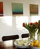 Obst und Blumen auf Esstisch dahinter drei abstrakte Gemälde an weißer Wand