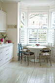 Moderne Küche mit weiß gestrichenem Boden und rundem Küchentisch unter großem Erkerfenster mit Fensterläden