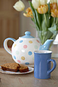 Frühstückstisch mit Teekanne, blauem Becher, Tulpen in einer Vase und einem Stück Kuchen auf einem kleinen Teller