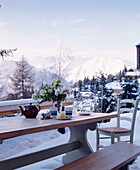Ein Outdoor-Detail von einem Holztisch und Sitzbank auf einer schneebedeckten Terrasse mit Blick auf die Berge in der Ferne gesetzt