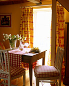 A country dining room yellow pattern curtains wood dining table with upholstered chairs in a red check fabric flower arrangement decanter and glasses